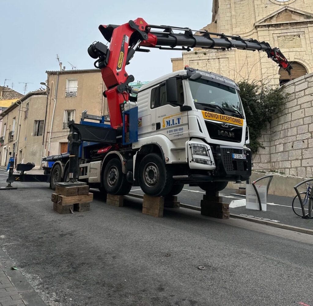 Camion-grue blanc M.L.T Palfinger sur cales en bois, CONVOI EXCEPTIONNEL, avec sa grue rouge étendue vers un bâtiment historique à horloge.