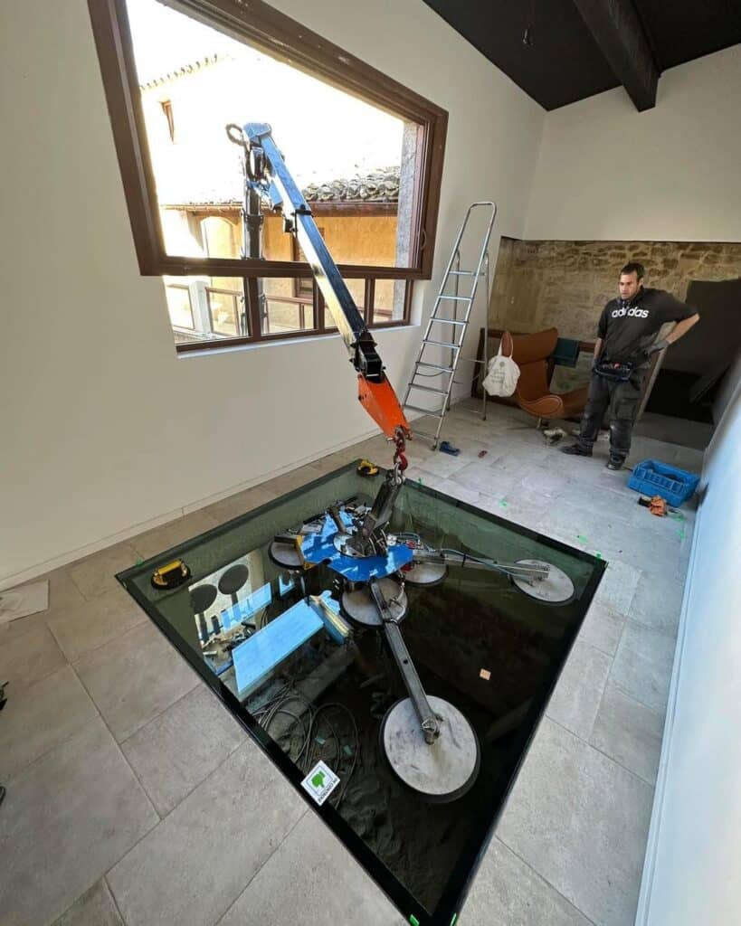 Grue robotisée positionnant une grande dalle de verre EN CONTROL pour un plancher vitré. Un homme en t-shirt Adidas supervise l'installation.