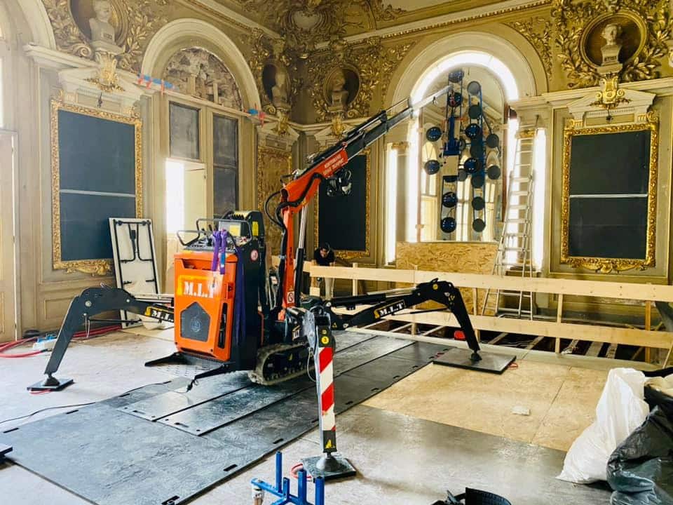 Mini-grue M.L.T. sur chenilles installant une grande vitre avec système à ventouses dans une salle dorée et ornementée d'un bâtiment historique.